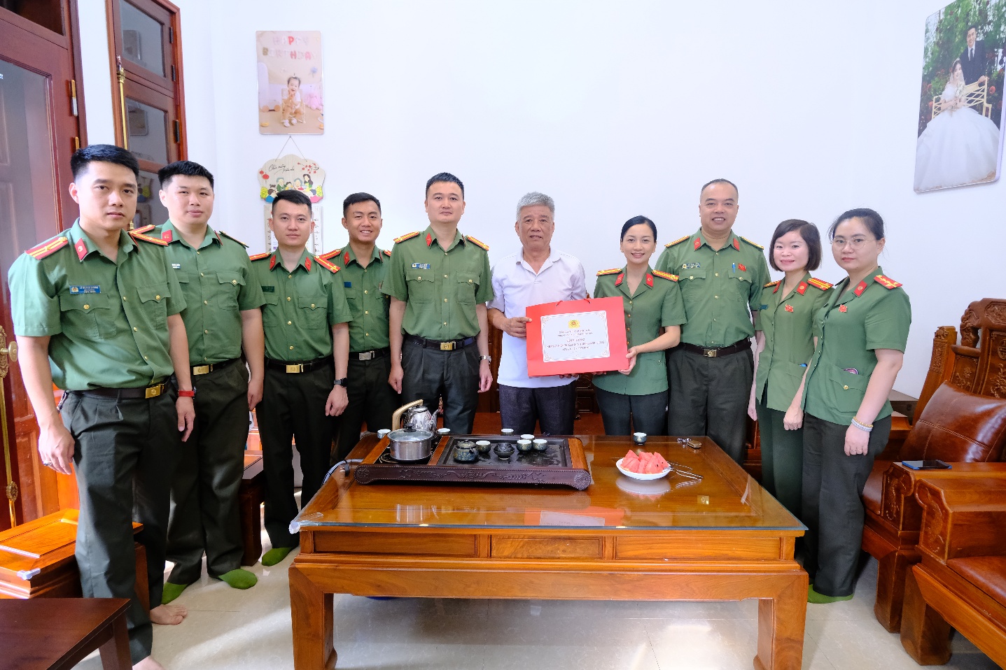 The image shows a group of people in green uniforms, seated around a table with a cake and a certificate, and a small table with decorations.

AI-generated content may be incorrect.